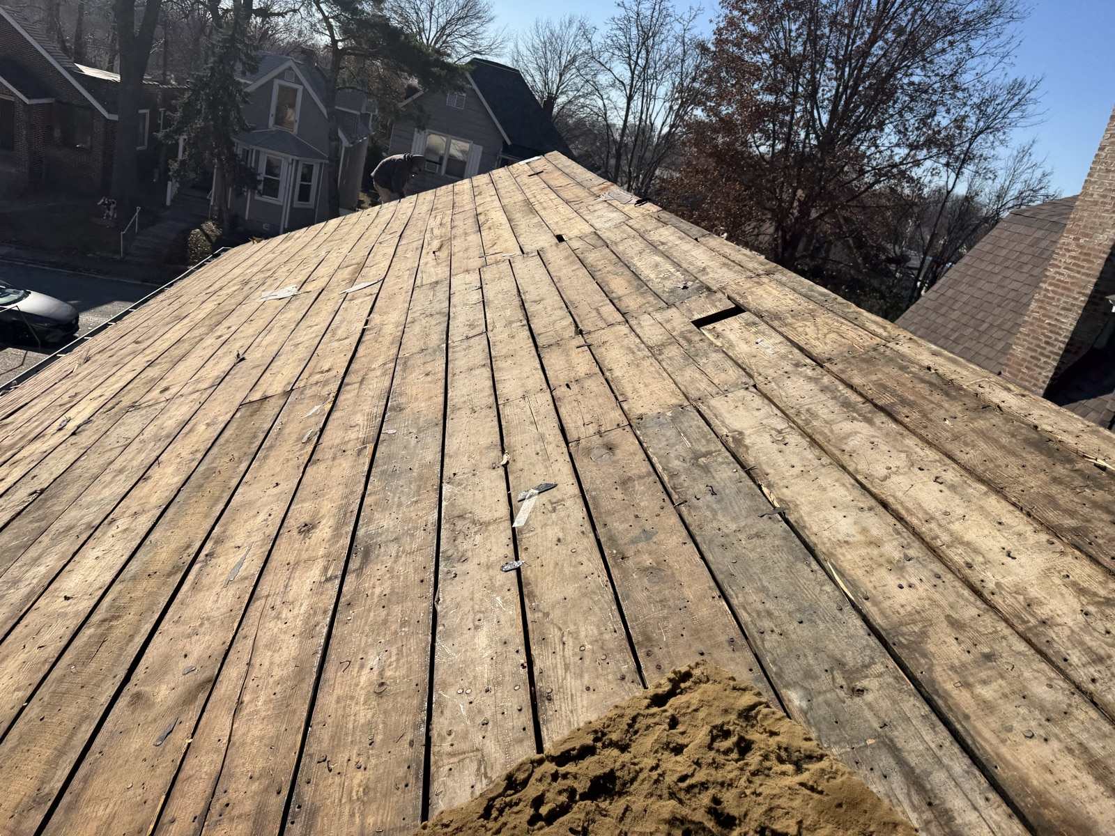 A roof plane stripped back to bare wooden plank decking studded with old nails, scraps of felt caught between the boards, a figure bent over at ground level beyond the roof beside a neighbouring grey house, and a street of two storey frame and brick houses under bare winter trees