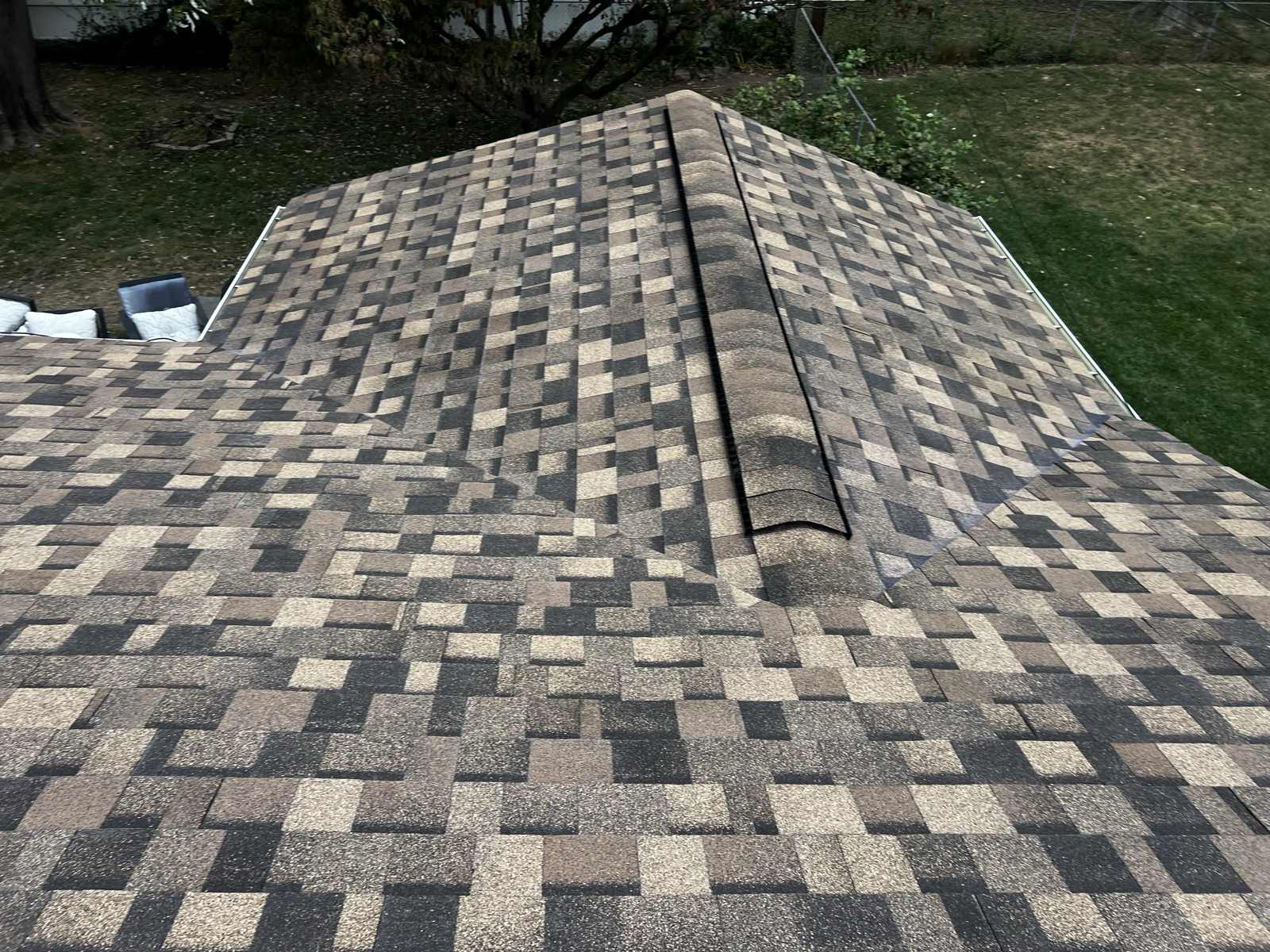 The hip of a newly laid brown, tan and charcoal multi tone architectural shingle roof capped with ridge cap shingles and carrying a dark ridge vent strip, the two planes falling away either side, with a mown lawn, chain link fence and a bare branched tree below