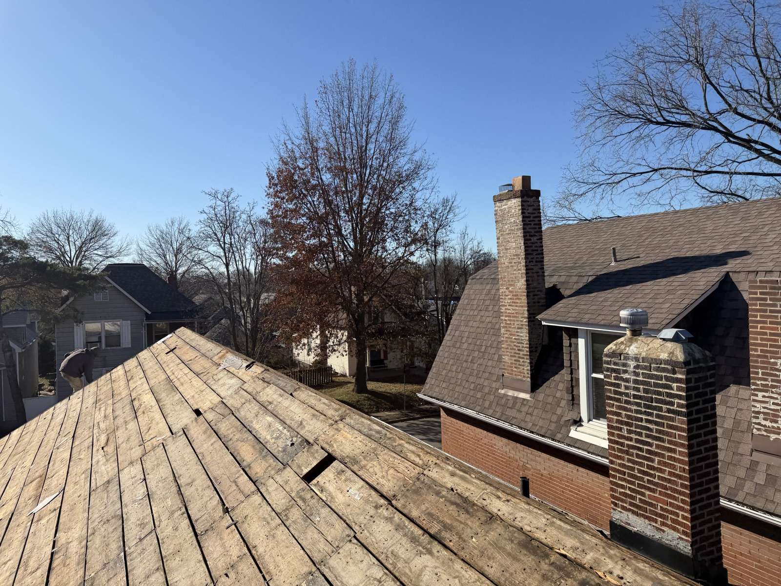 Stripped wooden plank decking across the lower left of the frame under a clear winter sky, bare and rust leaved trees in the middle distance, and at the right a red brick house still carrying its weathered brown shingles with two brick chimneys and a dormer vent