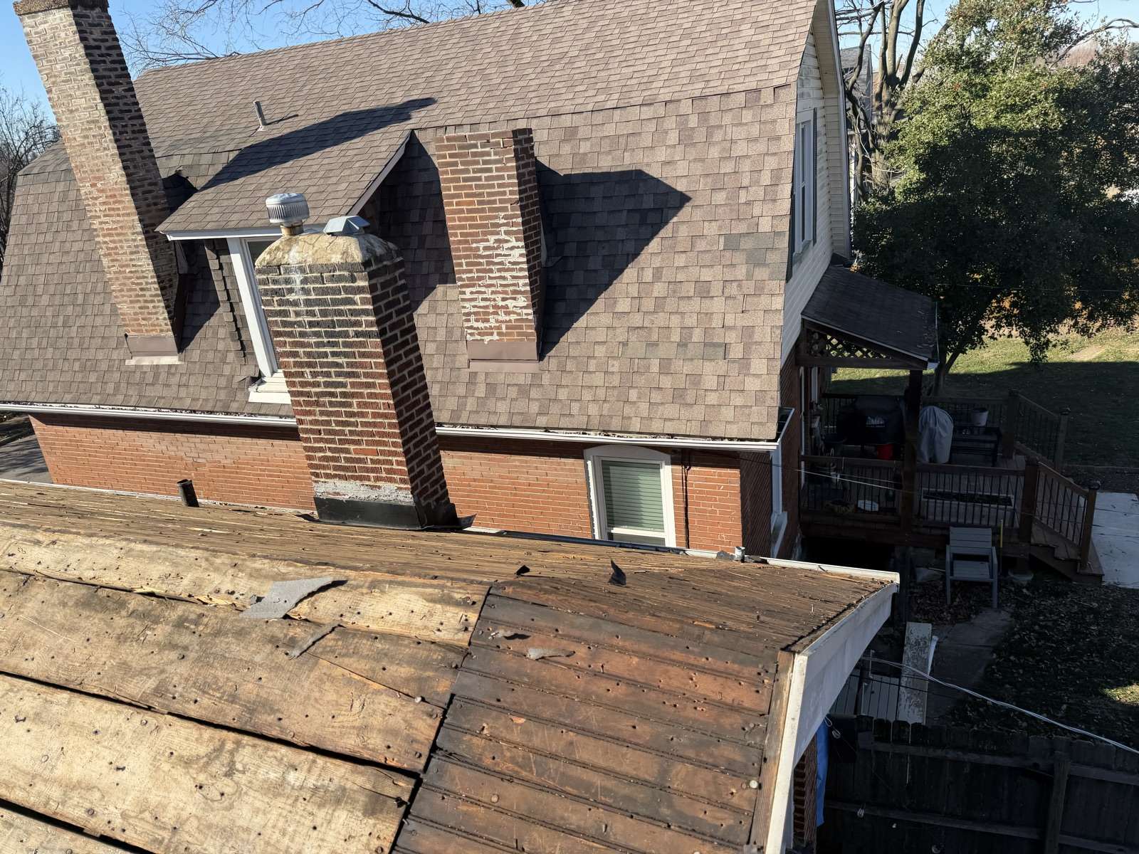 A lower roof plane stripped to dark weathered plank decking in the foreground while the two storey red brick house behind still carries its full weathered brown shingle roof, with two brick chimneys, a dormer with a metal turbine vent and a raised wooden deck at the right