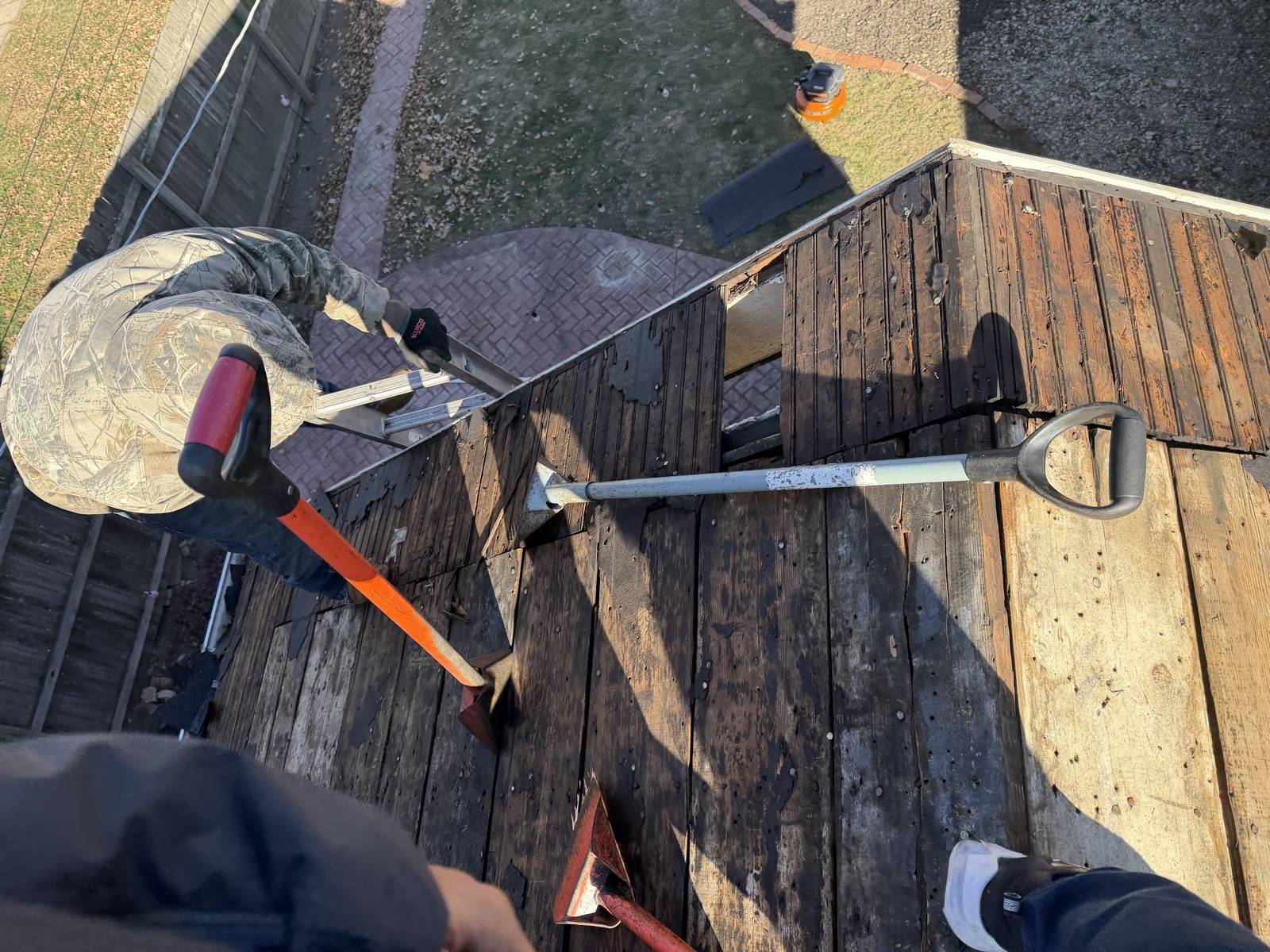 Looking down at stripped dark plank decking with tear-off tools laid across it, an orange handled tear-off bar, an aluminium D handle roofing shovel and two red flat spades, an aluminium ladder hooked over the eave and a worker in a camouflage jacket crouched at the roof edge, with a brick paver patio and lawn below
