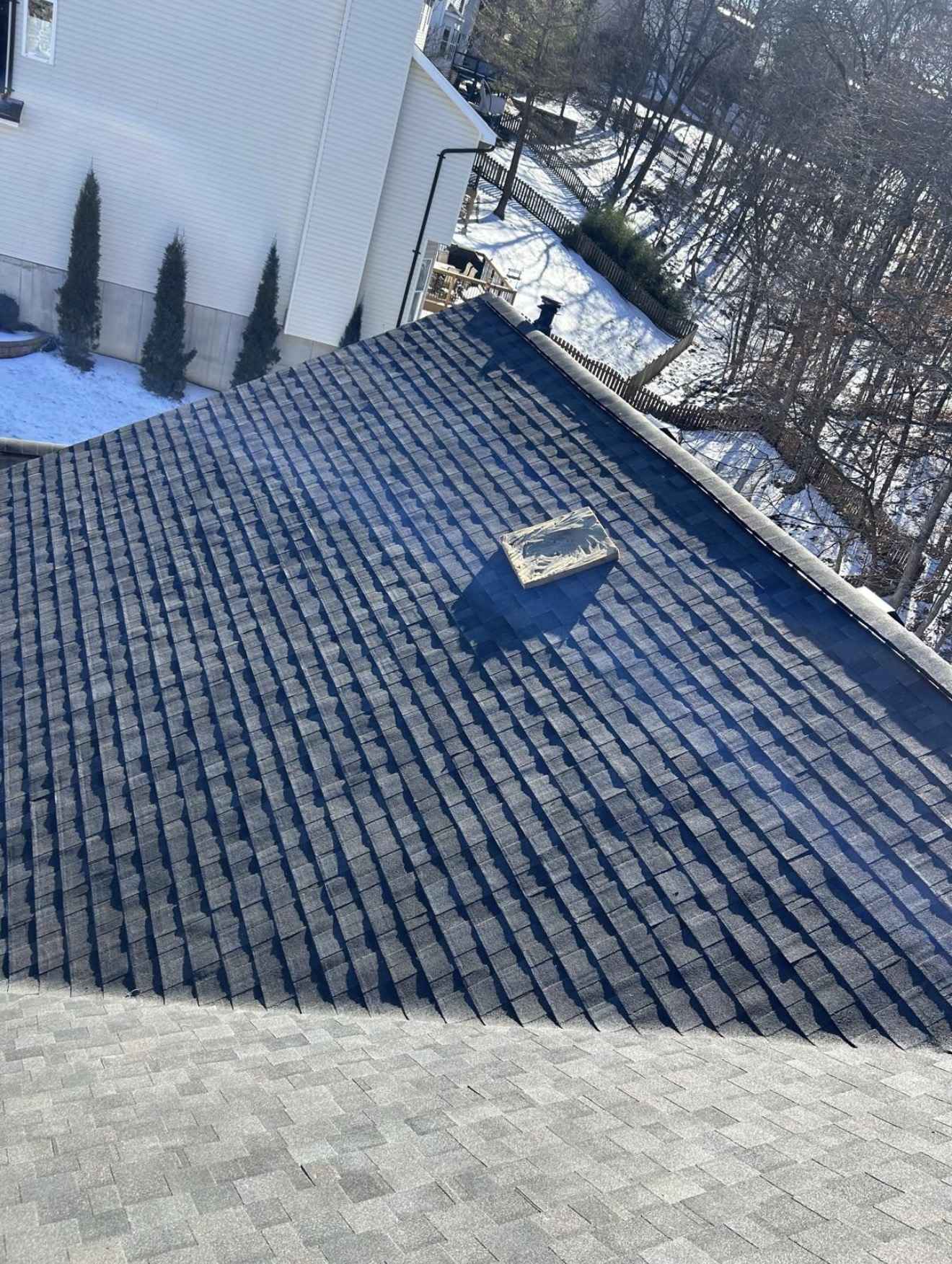 A roof mid installation, courses of dark charcoal architectural shingles laid down the upper plane meeting a lighter grey shingle surface across the lower third, an offcut of new shingle loose on the slope, with a cream vinyl sided neighbouring house, snow on the ground and bare winter trees beyond
